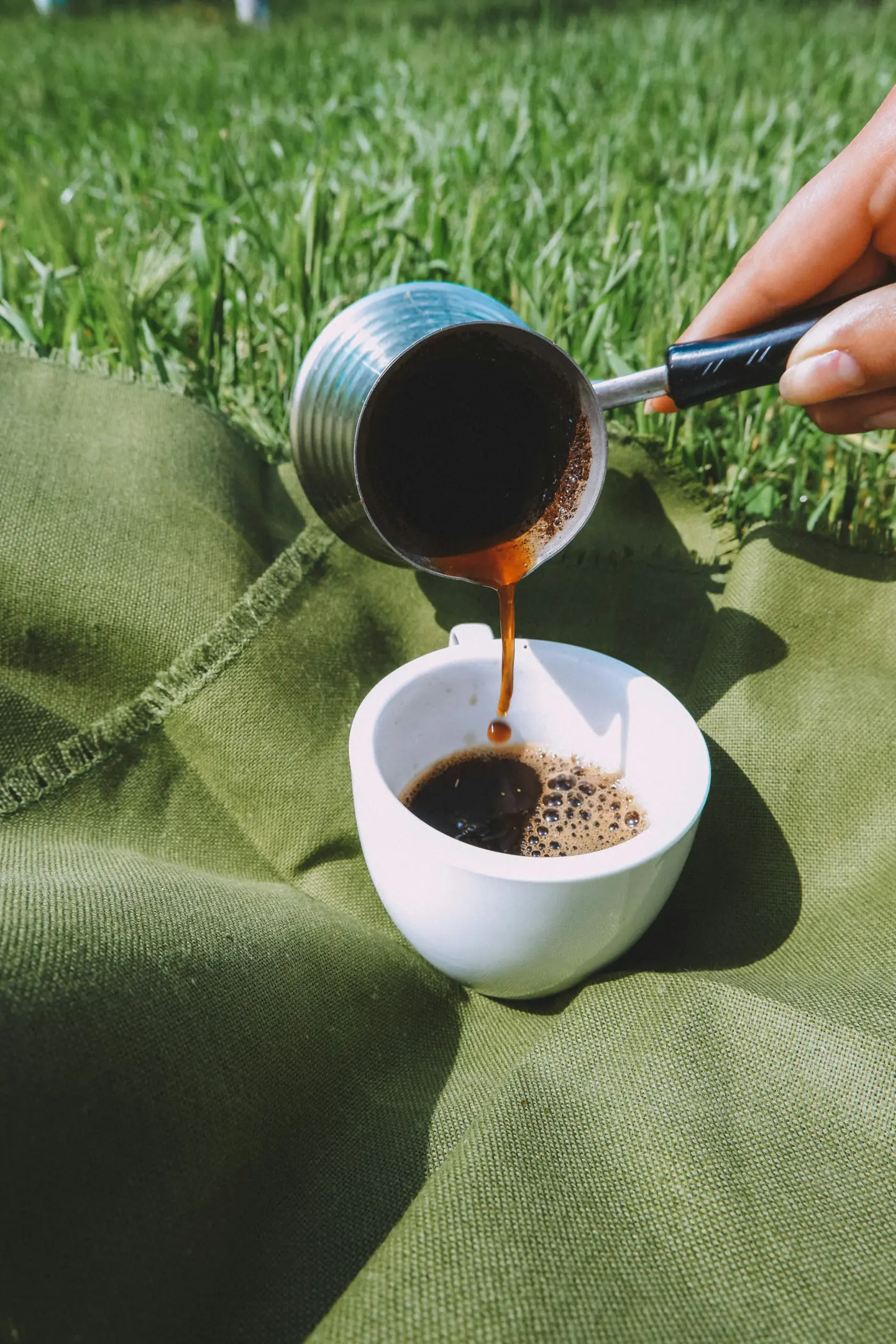 Café recién hecho vertiéndose en una taza blanca sobre una tela verde y hierba